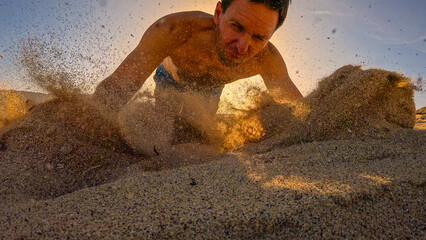 LOW ANGLE VIEW: Shirtless male runner trips over while running and falls onto sandy beach. Sand flies in all directions in golden morning light while young man hits the ground after spectacular fall.