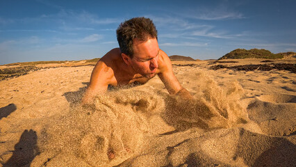 LOW ANGLE VIEW: Shirtless man falls forward onto the sand while running to the beach. Hands slam down and scatter sand into the air as late afternoon sunlight creates a dramatic, energetic atmosphere.