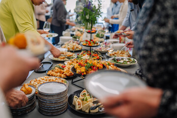 People gather around a buffet table filled with colorful appetizers, salads, and snacks during a catered event, serving themselves with plates in hand.