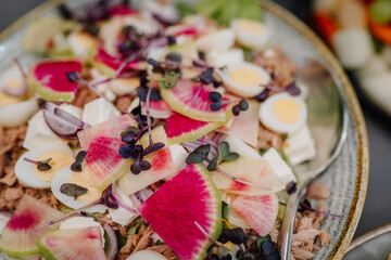 Close-up of a vibrant salad featuring sliced watermelon radish, boiled eggs, feta, microgreens, and tuna served on a ceramic plate with a spoon.