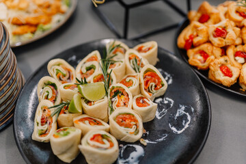 Close-up of neatly arranged tortilla roll-ups filled with vegetables and herbs, garnished with lime and rosemary on a black serving plate at a buffet.