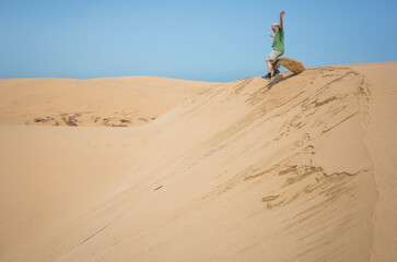 Man jumps from a tall sand dune near Essaouira, Morocco, leaving a trailing plume of sand behind him. Bright blue sky and golden dunes highlight the sense of movement and freedom