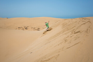 Man running fast down a steep sand dune near Essaouira, Morocco, kicking up clouds of sand with each step. Dynamic action against golden dunes and clear blue sky on the Atlantic coast