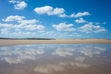 Clouds reflected in wet sand on the beach in Essaouira, Morocco. Low tide creates a mirror-like surface under a vibrant blue sky with scattered white clouds