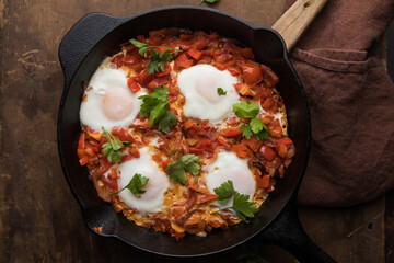 Shakshuka in a frying pan on a dark wooden background. Eggs cooked in tomato sauce with spices and fresh parsley on top. Rustic style. Top view,