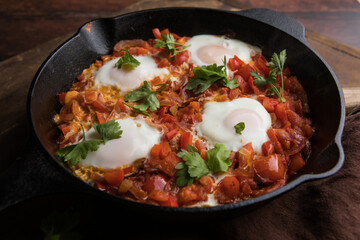 Shakshuka in a frying pan on a dark wooden background. Eggs cooked in tomato sauce with spices and fresh parsley on top. Rustic style. Top view,