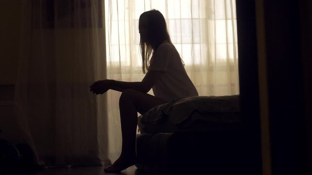 A woman sits alone on the edge of a bed in an empty room, her silhouette framed by soft light. She appears lost in thought, emotionally overwhelmed, facing a personal mental health crisis.