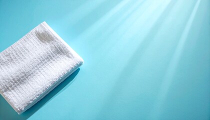 淡いマットブルーの背景上に白いビーチタオルの折り目がきれいに見え、柔らかい自然光が生地の細かな凹凸を繊細に照らす

