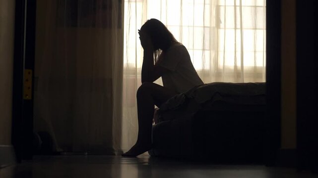 A woman sits alone on the edge of a bed in an empty room, her silhouette framed by soft light. She appears lost in thought, emotionally overwhelmed, facing a personal mental health crisis.
