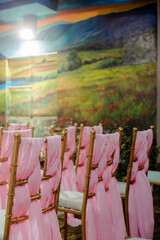 Rows of gold chairs adorned with pink fabric sashes, creating an elegant and celebratory atmosphere against a backdrop of a painted landscape scene.
