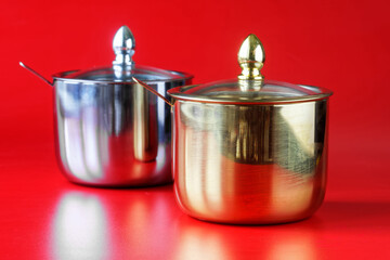 Two metallic sugar bowls—one silver, one gold—each with a lid and spoon set against a vibrant red background, ideal for kitchenware catalogs or culinary blog imagery. Photo