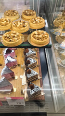 A display of assorted desserts in a bakery. The scene includes round nut-topped cakes and triangular chocolate slices with white icing.