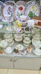 A display case filled with decorative porcelain teacups, plates, and figurines. The items feature floral designs and vibrant colors, showcasing fine craftsmanship.
