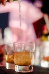 Glass filled with a spirit and mixer, showcasing a mixed beverage and nightlife setting with pink hues and bartender pouring a clear liquid in soft focus.