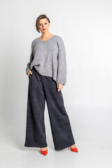 Portrait beautiful woman of model appearance. Girl in grey jumper, black trousers, red shoes on grey background. Pretty female with trendy makeup posing in studio. Vertical photo.