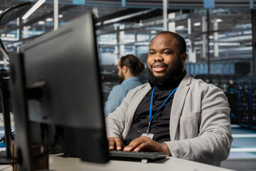 Cheerful data center employee working to update rackmounts software, configuring network virtualization. Upbeat african american man in server room verifying mainframes settings