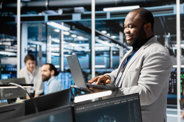 Happy data center facility admin using laptop to update supercomputers software, configuring network virtualization. Jolly server hub IT professional using notebook to verify equipment settings