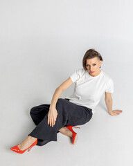 Portrait beautiful woman of model appearance. Girl in white T-shirt, black trousers, red shoes on grey background. Pretty female with trendy makeup posing in studio. Vertical photo.