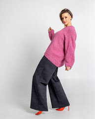 Portrait beautiful woman of model appearance. Girl in pink jumper, black trousers, red shoes on grey background. Pretty female with trendy makeup posing in studio. Vertical photo.