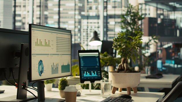 Modern workspace with analytical data dashboards displayed on devices in bright office environment. PC screen on desk with financial statistics graphs next to potted plants, zoom in shot