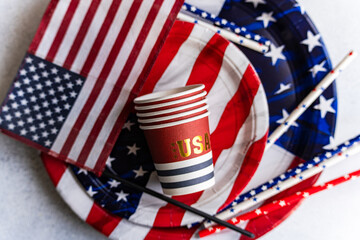 Overhead view of a stack of paper cups, paper plates, drinking straws and an american flag on a table for Independence Day on fourth of July