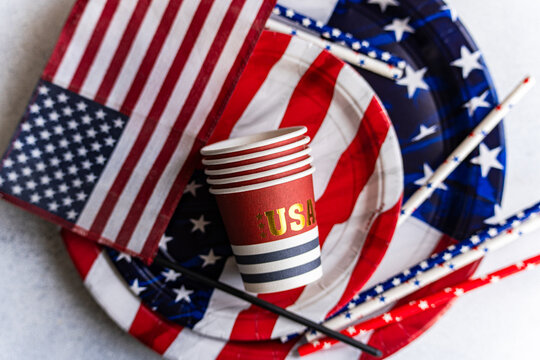 Overhead view of a stack of paper cups, paper plates, drinking straws and an american flag on a table for Independence Day on fourth of July