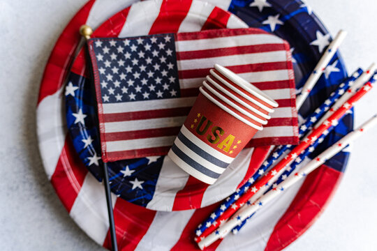 Overhead view of a stack of paper cups, paper plates, drinking straws and an american flag on a table for Independence Day on fourth of July