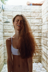 young woman with long hair taking outdoor shower in sunlight wearing white bikini top, expressing relaxation and refreshment, stone wall background