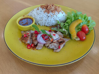 thai food, waterfall pork, fried garlic on rice and nam jim jaew in a yellow bowl, green vegetables, 1 piece of pumpkin, 2 tomatoes on a large yellow dish on a wooden background, food, copy space