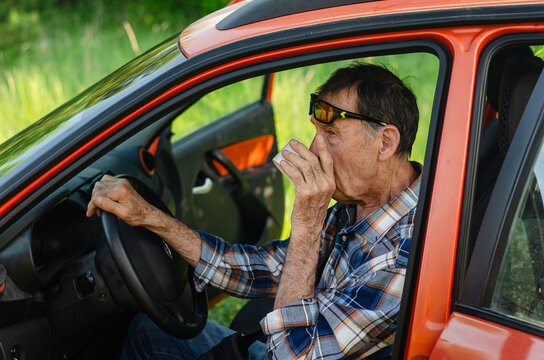 Senior man sipping tea in orange car, hand covering mouth. Sunglasses perched on forehead, relaxed grip on wheel. Open doors frame lush bokeh grass. Warm golden hour atmosphere
