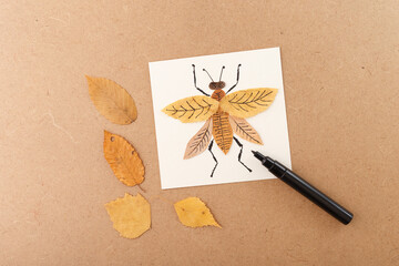 A creative insect illustration made using a dried leaf for the body and black marker for details. The artwork is placed on white paper with additional leaves on a brown background