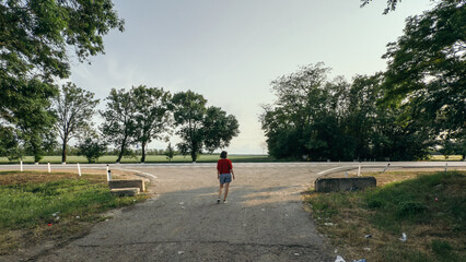 Woman walking alone on an empty rural road toward a tree-lined horizon in the evening light, surrounded by greenery and silence, solitude concept