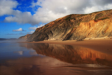 Portugal, Algarve Region, Vila do Bispo, South-West Alentejo and Vicentine Coast Natural Park. Panoramic view of the huge cliffs reflected on the wet sand of a deserted Vale Figueiras Beach.