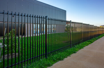 A black vertical metal security fence lines a path in front of a contemporary institutional or...