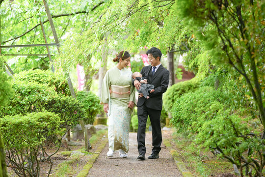 On a spring day in May in a Japanese garden, a one-month-old Japanese baby in hakama is held by a suited man, walking with a kimono-clad mother, both in their twenties, sharing a gentle family walk.