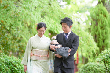 On a spring day in May in a Japanese garden, a one-month-old Japanese baby in hakama is held by a suited man, walking with a kimono-clad mother, both in their twenties, sharing a gentle family walk.