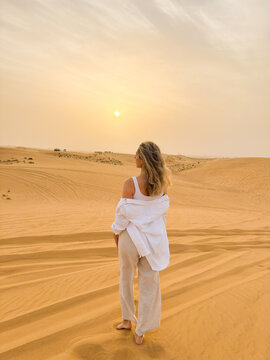 Woman standing in the desert, looking at the setting sun, facing backwards to the camera.