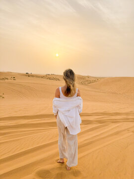 Woman walking on sand dunes in the desert towards the setting sun, facing backwards to the camera.