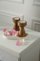Elegant candlesticks with burning candles and flower petals on dresser near white wall, closeup