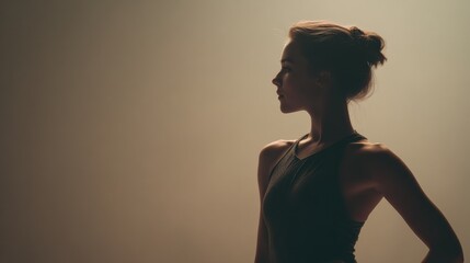 Profile of a young graceful ballerina posing with hand on hip, illuminated by soft light against a neutral background, creating a serene and artistic atmosphere