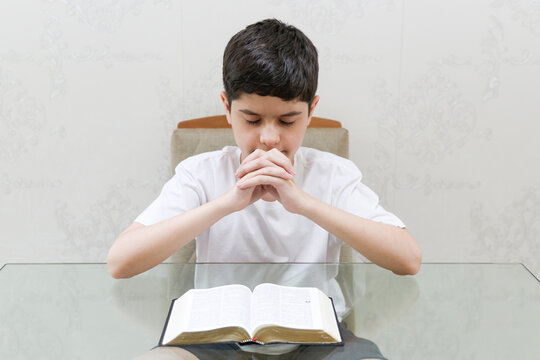 Brazilian preteen boy prays quietly with his hands clasped and head bowed in front of an open Bible. Eyes closed, he wears a white shirt and sits at a table indoors