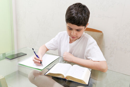 Brazilian preteen boy reads the Bible and writes in his notebook while studying at home. He is right-handed, wears a white shirt, and sits at a table indoors