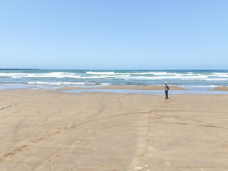 On a breezy spring day in May by the Sea of Japan, a Japanese man in his late seventies walks alone on the beach, captured from above in an aerial photo showing waves and quiet solitude.