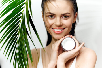 Smiling model holding skincare cream with tropical greenery background, showcasing beauty and freshness in wellness concept.