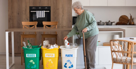 Senior woman throwing plastic bottle into garbage bin in kitchen. Waste sorting concept