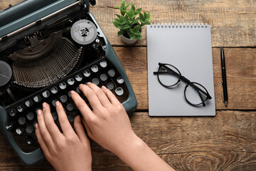Woman typing on typewriter with eyeglasses, notebook and houseplant on brown wooden background