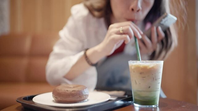 Close up of an iced matcha latte and chocolate donut on a cafe table with a woman in the background.