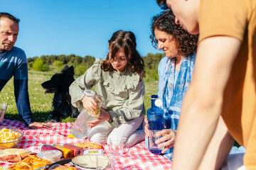 Happy family having fun, eating and playing with their dog during a picnic in a park, girl is pouring lemonade