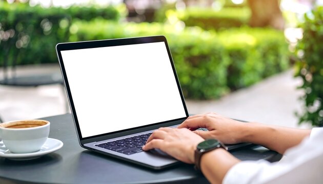Person typing on a laptop with blank white screen, seated at outdoor table next to coffee cup.