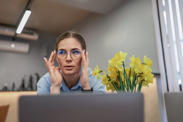 Woman Glasses Laptop: Focused worker adjusts glasses near daffodils at office desk during day.